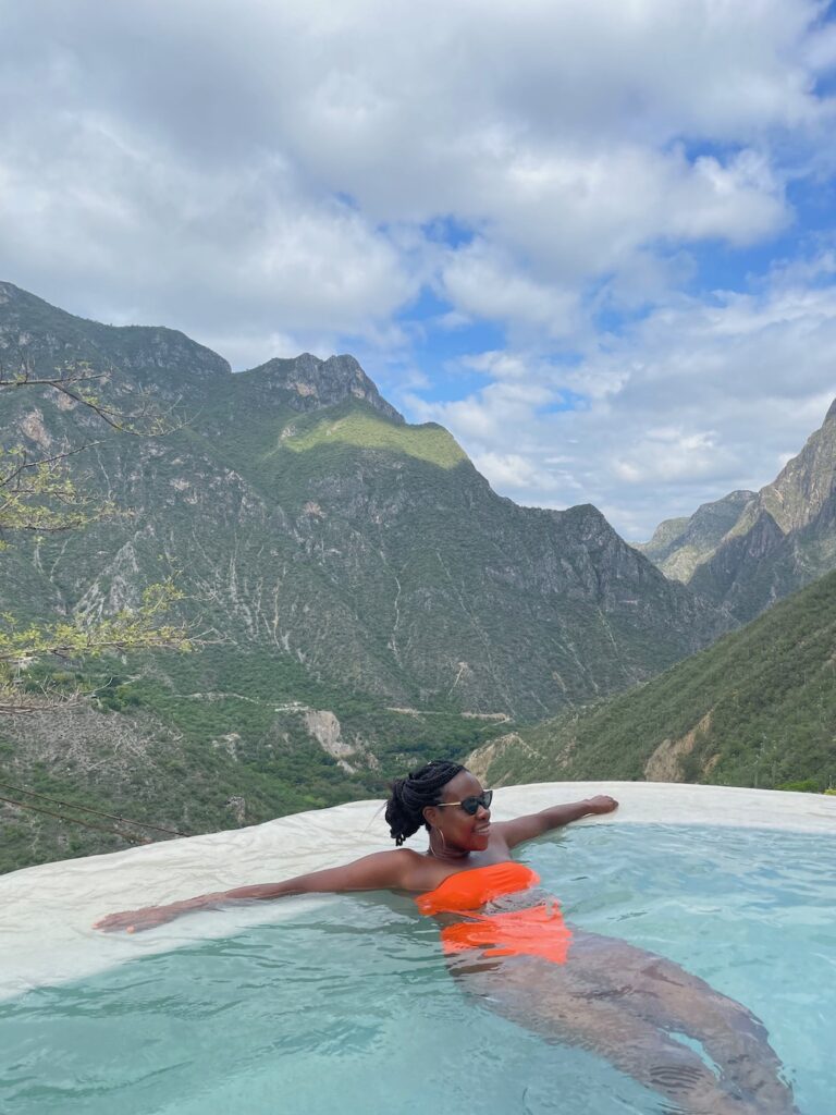 Pretty Black lady wearing an orange bathing suit sitting in a natural pool surrounded by mountains.