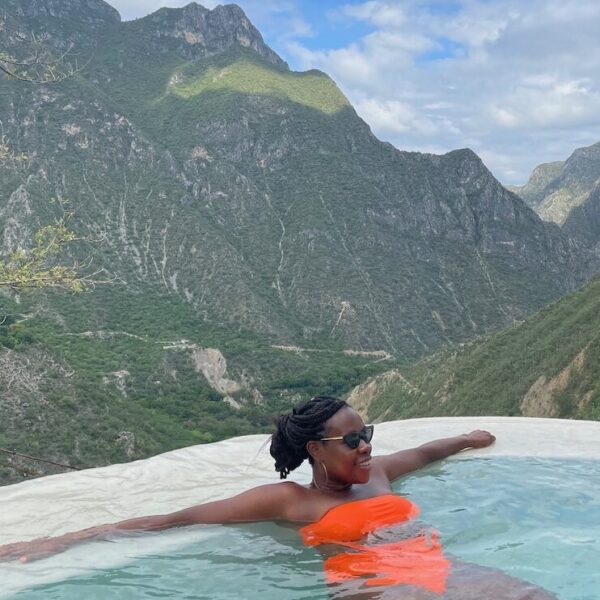 Pretty Black lady wearing an orange bathing suit sitting in a natural pool surrounded by mountains.