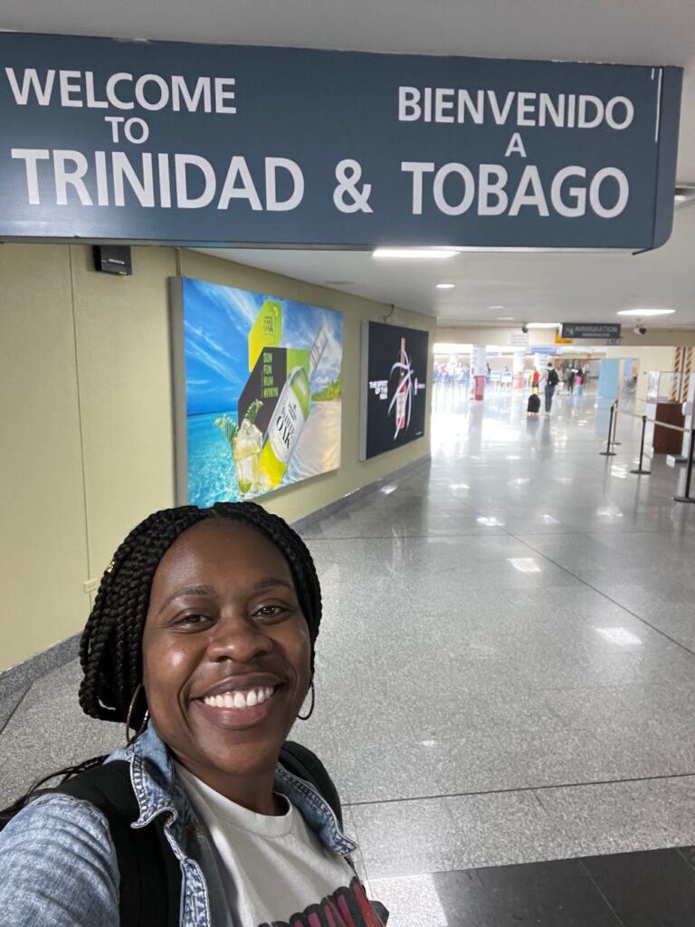 Black woman standing in front of Trinidad and Tobago welcome sign in airport.  Trinidad Carnival 2024 planning guide.