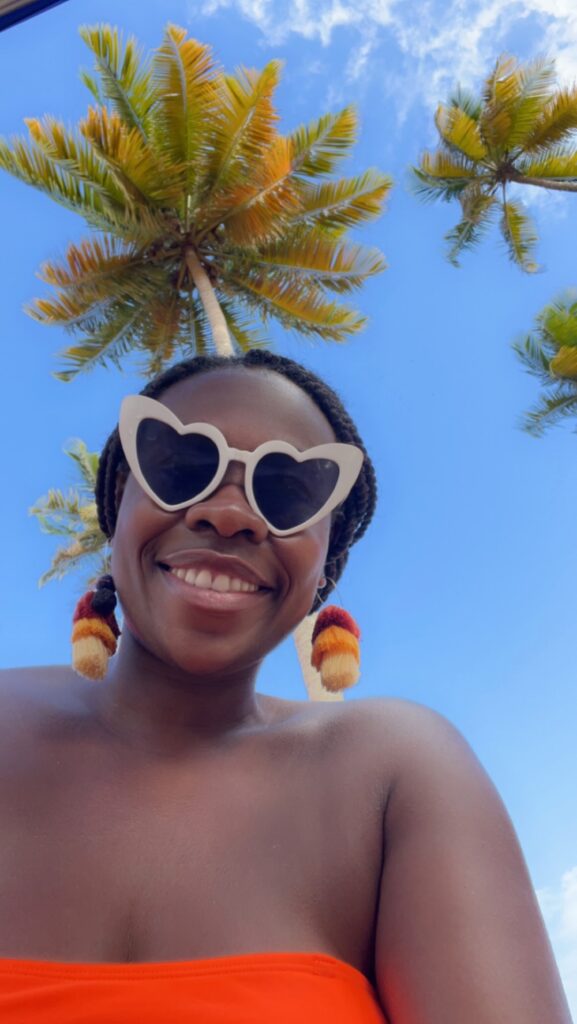 Black lady poising in an orange bathing suit with palm trees above her.