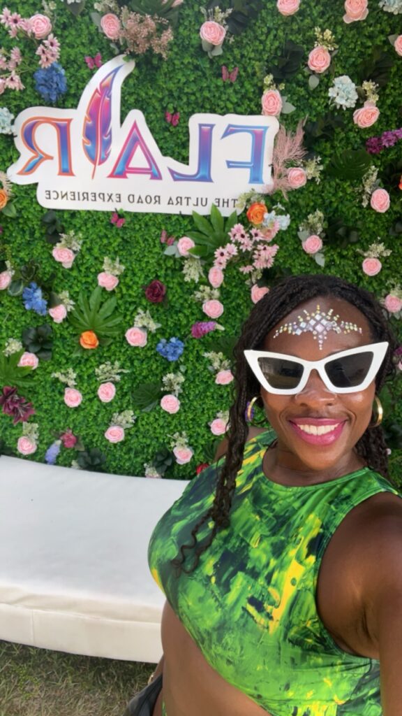 Black lady wearing a green bathing suit with white sunglasses standing in front of a  green wall covered in pink, orange, purple and blue flowers with a sign that reads FLAIR the ultimate road experience. 