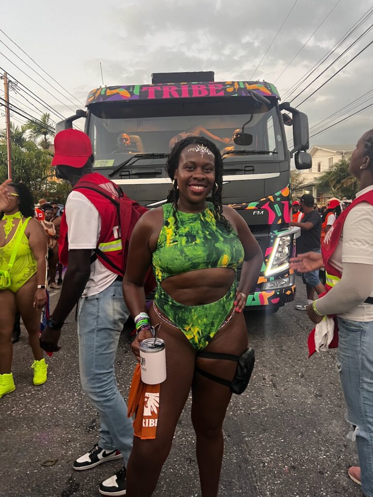 Black ladying wearing a green bathing suit with a black leg bag, holding a cup smiling in front of a truck on the road that reads TRIBE.  A man dressed in a red vest and jeans is standing behind her in Port of Spain, Trinidad. 