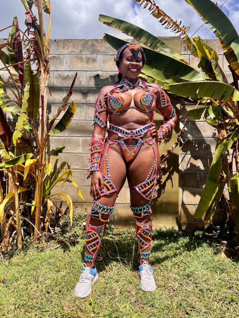 Pretty Black Lady dressed in a colorful costume posing on grass behind lush leaves in Port of Spain for Trinidad Carnival.