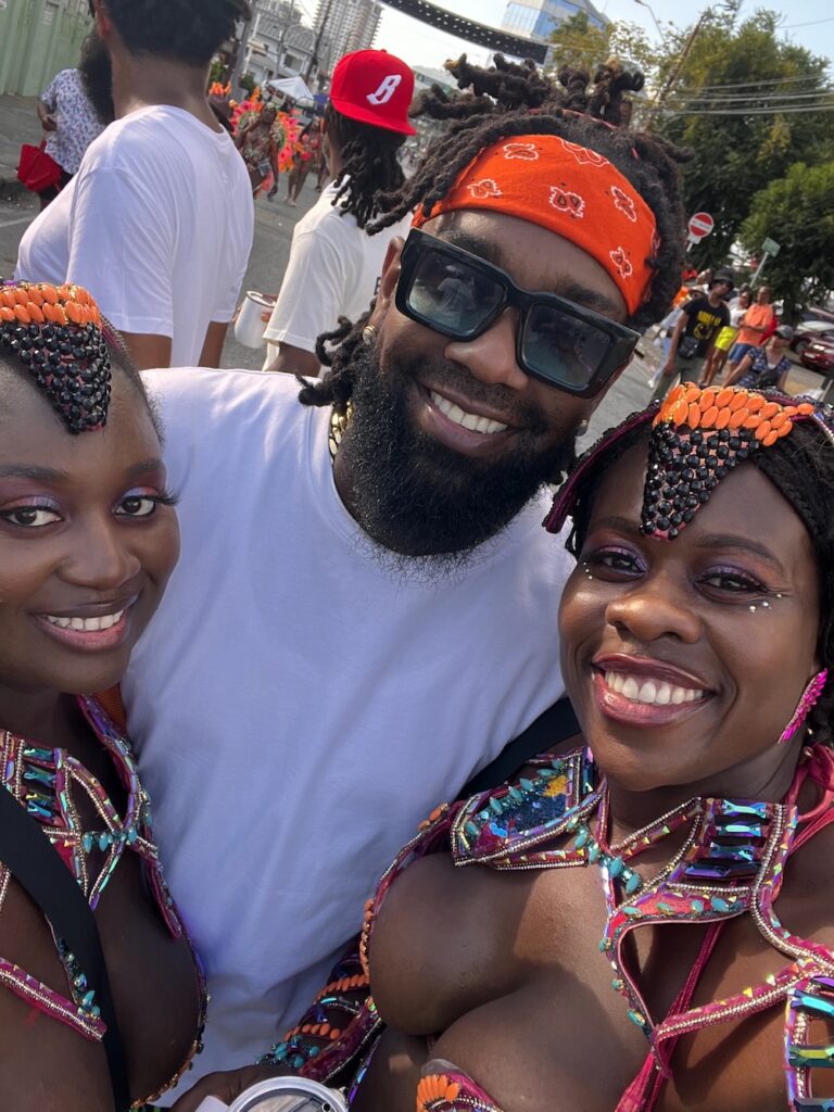 2 beautiful Black masqueraders dressed in colorful costumes for Trinidad carnival smiling and posing for a picture with the soca artist Lyrikal. 