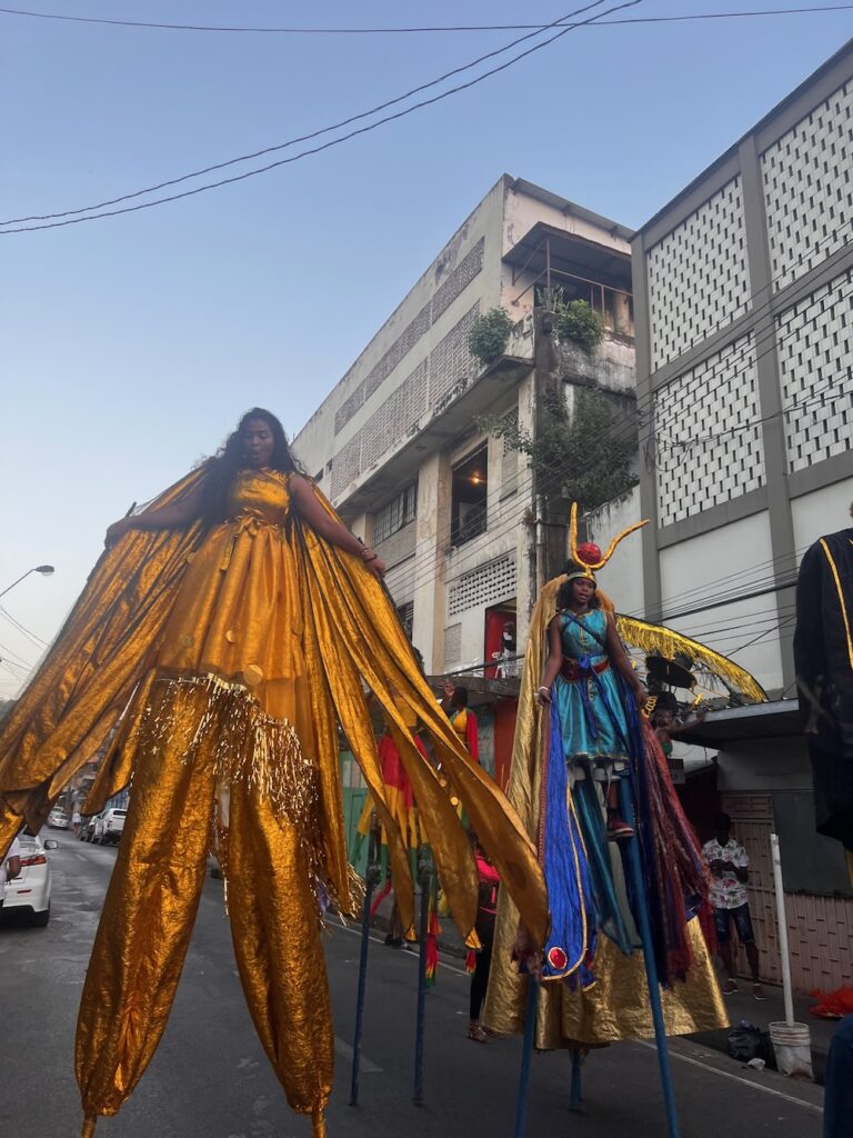 A beautiful woman dressed in a marigold and jewel costume on stilts. Another masquerader on stilts in the background dressed in blue and gold costume.  