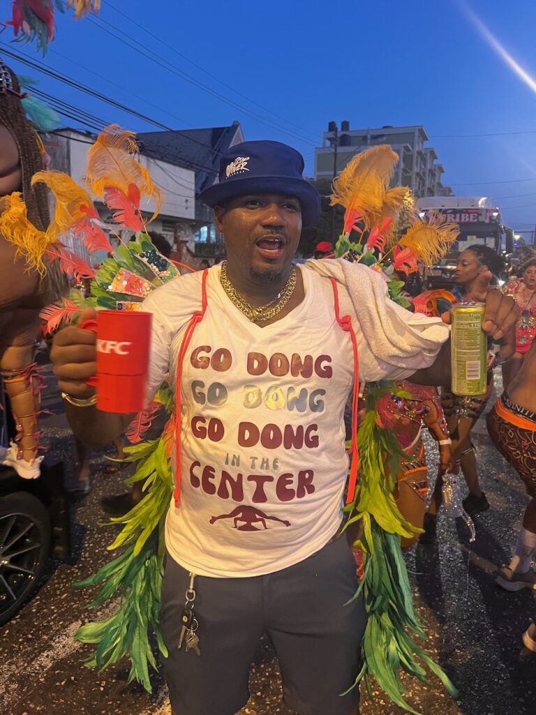 A man holding up cup enjoying the Trinidad Carnival parade with a shirt that says Go Dong, Go Dong Go Gond, in the Center. 