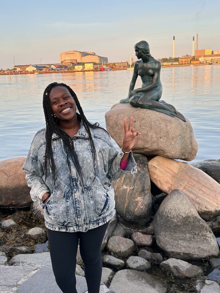 Black woman wearing a jean jacket standing smiling with a peace sign front of little a mermaid statue in Copenhagen, Denmark.  15 Tips for how travel with a Full-Time Job. 