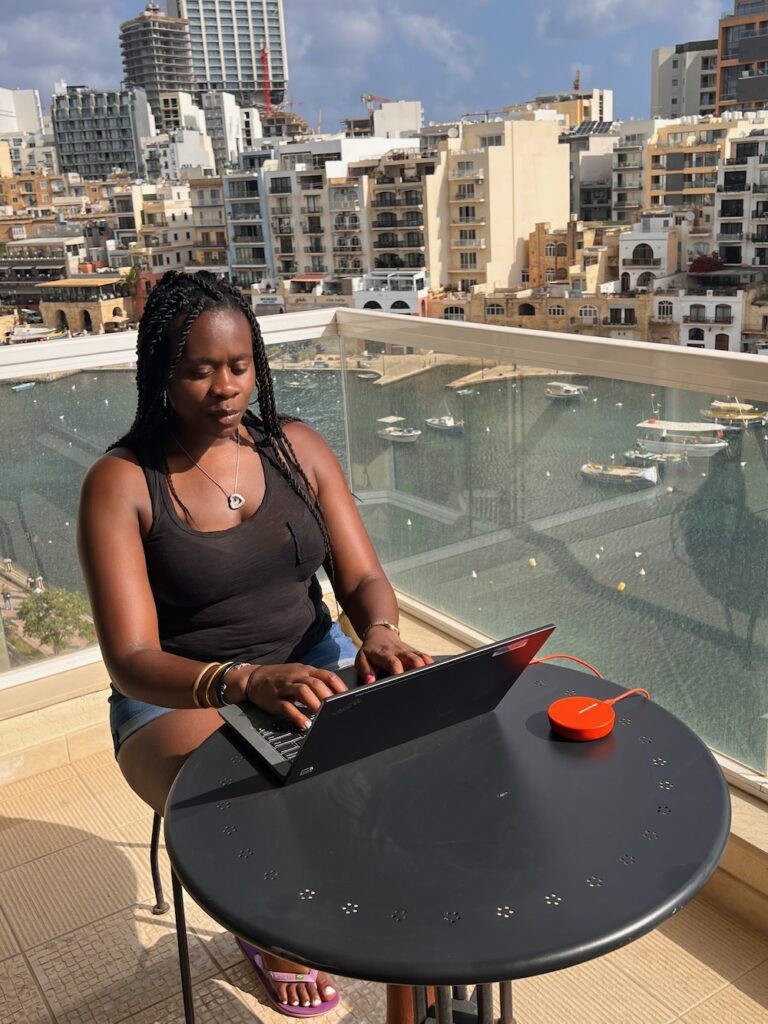 Black lady sitting on a balcony in St. Julian's Malta working on laptop and portable hot spot/. 15 Ways to Travel with a Full-Time Job.