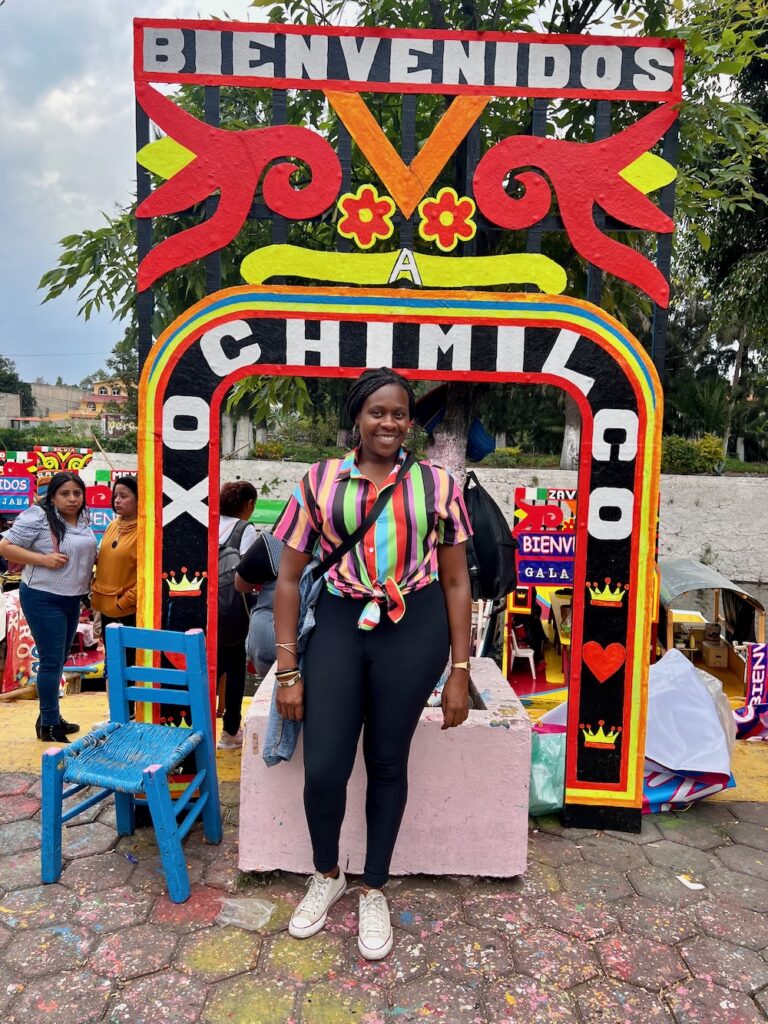 Black women in a colorful shirt with black pants smiling in front of the Xochimilco and bienvendio signs in Mexico City. 