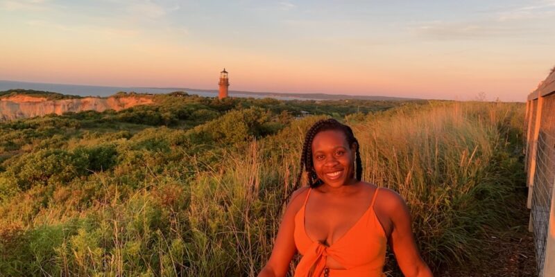 Black lady smiling in an orange dress with lighthouse and cliff in the background.