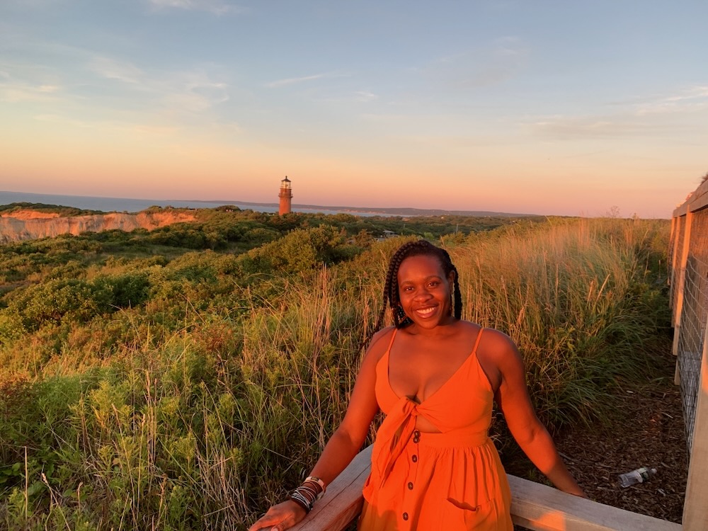 Black lady smiling in an orange dress with lighthouse and cliff in the background.