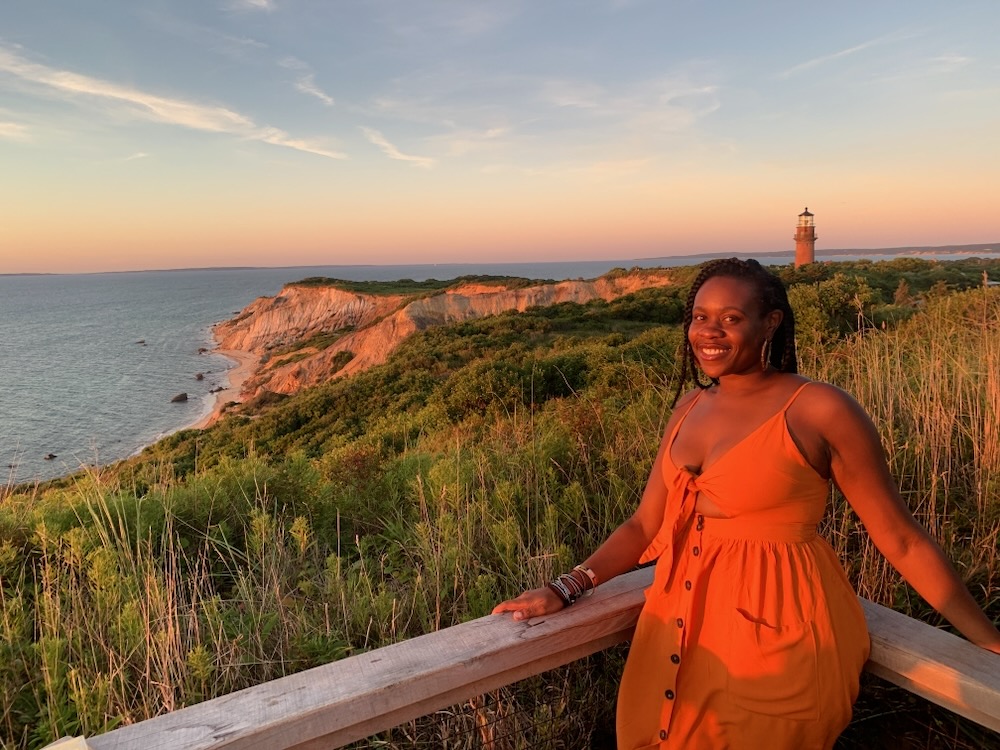 Black woman wearing an orange dress smiling with cliffs, lighthouse and sunset in the background in Aquinnah, Martha's Vineyard. 