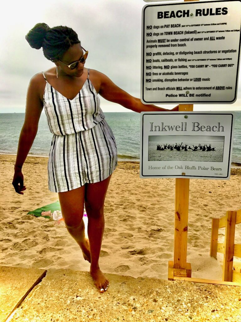 Black lady wearing a black and white coverup with sunglasses looking away from the camera but standing next to the Inkwell beach sign and beach rules. 