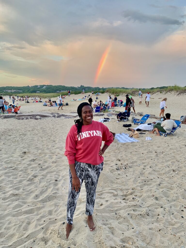 Black lady smiling and posing on the beach with double rainbows and people in the background