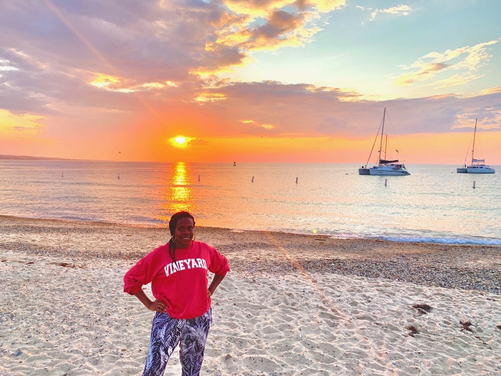 Black lady smiling with hands on her hips with sunset and sailboats in the background