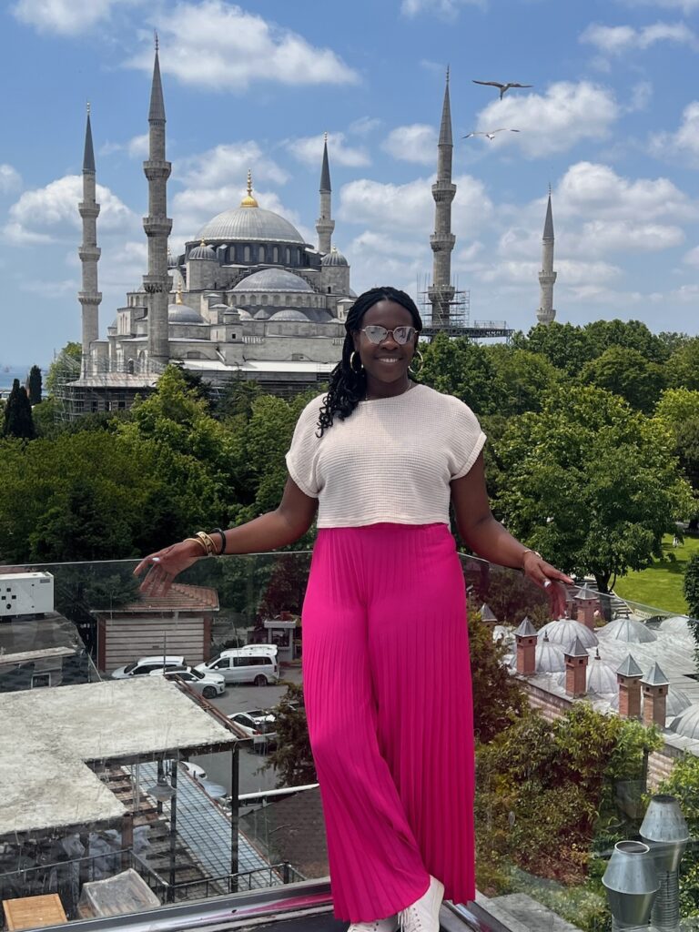 Black lady wearing pink pants, white shirt smiling in front of the Blue Mosque in Istanbul, Turkey with seagulls flying above.  Istanbul for first time visitors