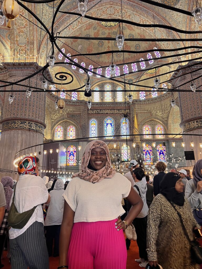 Black lady smiling with pink pants, white shirt and scarf covering her head.  She is posing inside the Blue Mosque in Istanbul, Turkey. 