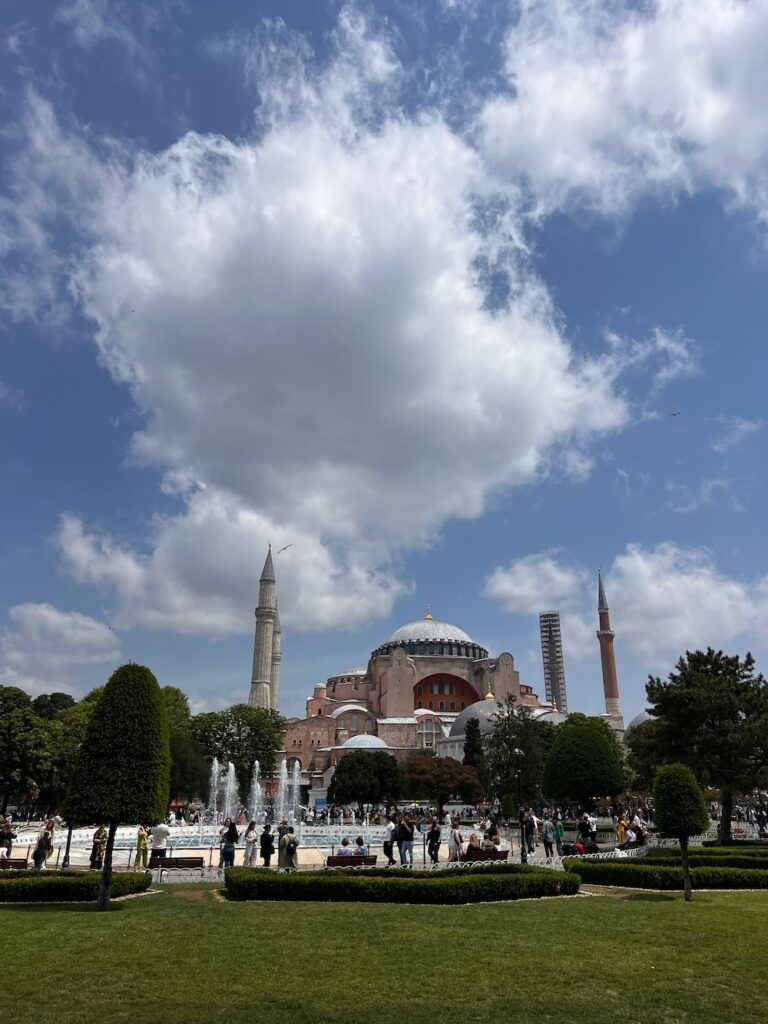 Hagia Sophia grand mosque on a party sunny day.  In front of the fountain with tourists standing nearby.
