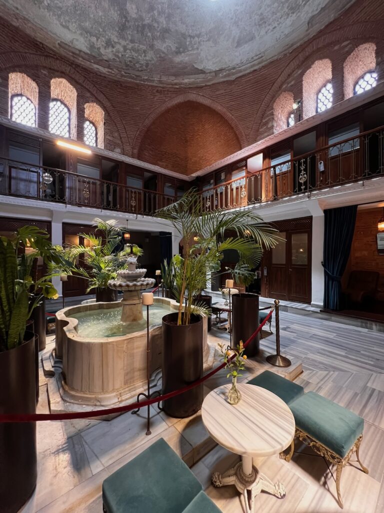 A lush fountain surrounded by green seats and a marble colored table with green plants Inside the Cagaloglu Hamami, Turkish bathhouse in Istanbul. 