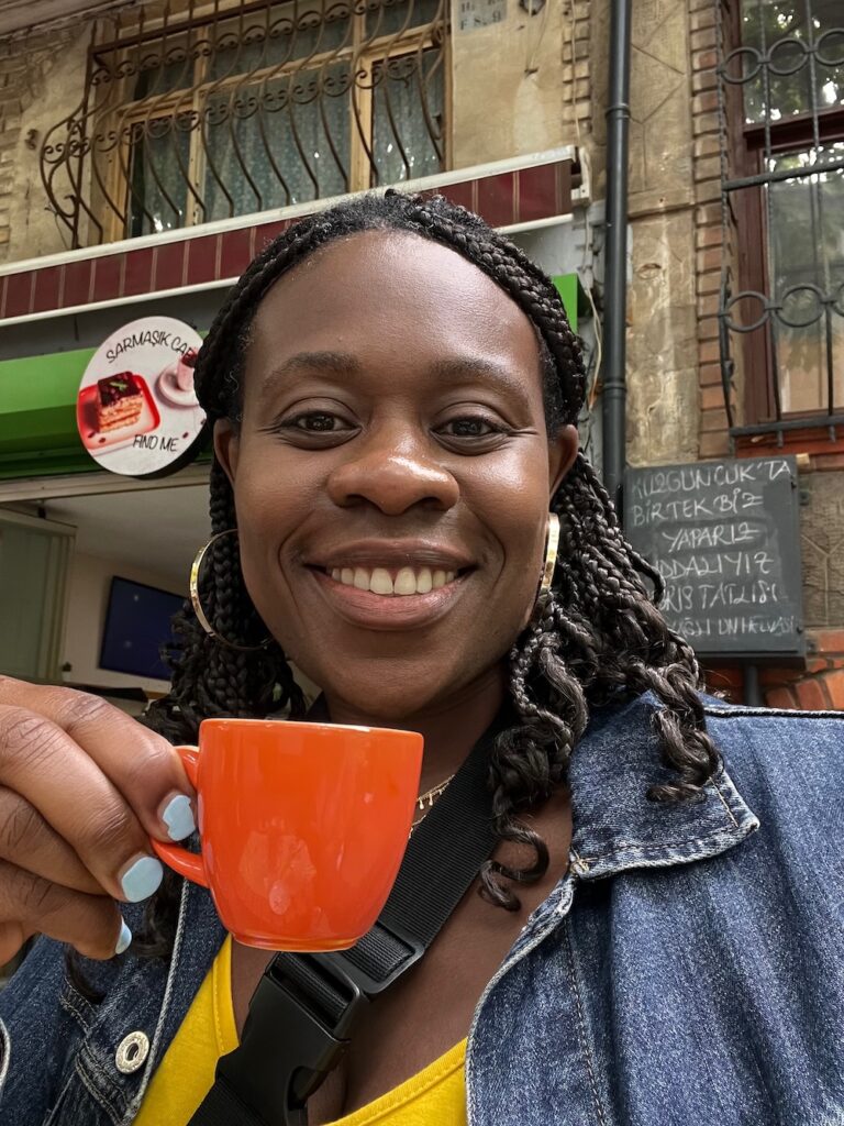 Black lady smiling holding a red coffee cup wearing a blue jean jacket, yellow dress and gold hoop earrings. 