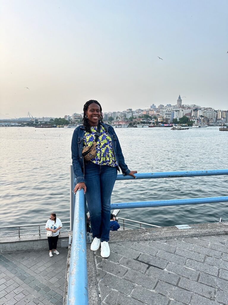 Black lady smiling wearing jean jacket, colorful shirt, blue jeans and white shoes on the Galata Bridge in Istanbul, Turkey. What to Pack for Istanbul. 