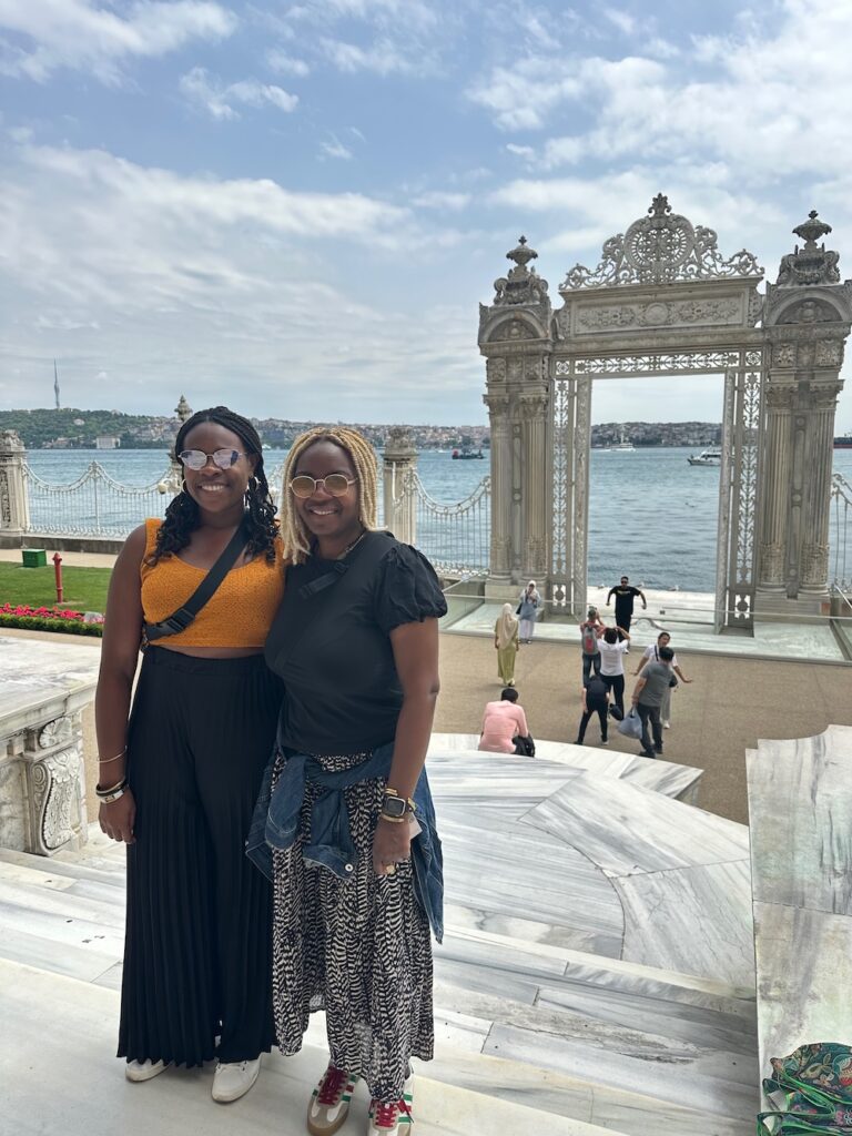 2 Black ladies standing in front of the gate at Tokapi palace in Istanbul.  Lady on the right is wearing black shirt with flowy black and white pants. The lady on the left is wearing an orange top and black pants. 