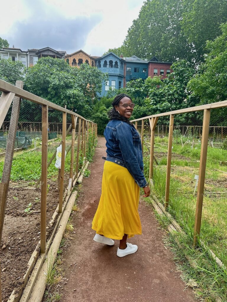 Black lady smiling wearing a yellow dress, jean jacket, white sneakers. at the Bostan Garden in Kadikoy, Turkey.  What to pack for Istanbul