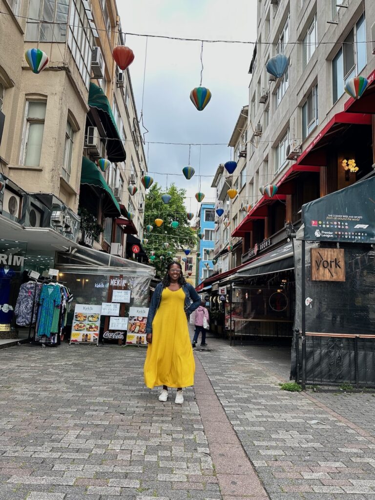 Black lady smiling wearing a beatufil yellow dress, jean jacket and white sneakers standing in the middle of a street in Istanbul, Turkey. 