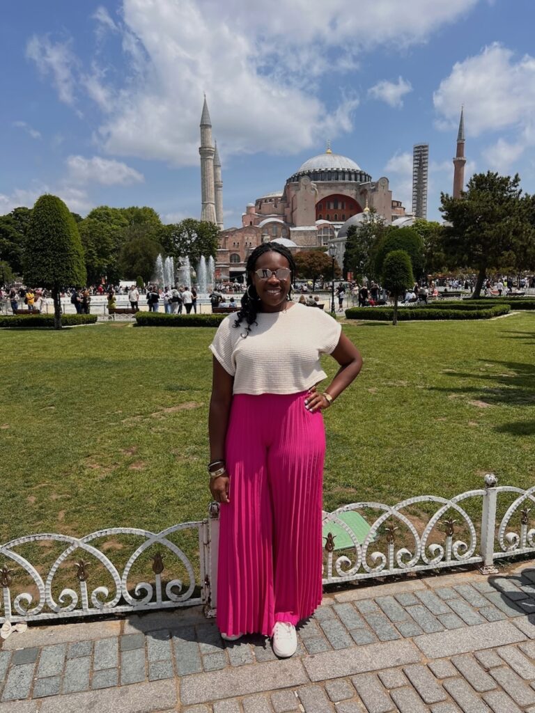 Black lady standing and smiling in front of Hagia Sophia wearing sunglasses, white shirt and pink pants.  What to pack for Istanbul. 