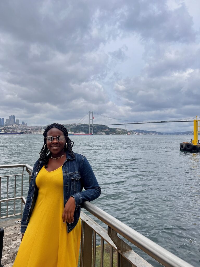 Black lady wearing a yellow dress with a blue jean jacket, wearing sunglass and smiling in front of the Bosphorus touring the Asian side of Istanbul.